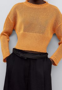 Strickpullover in leuchtendem Orange, geschnittenes Design mit geripptem Saum und Rundhalsausschnitt. Kombiniert mit schwarzen, plissierten Hosen und einem gestreiften Bund.