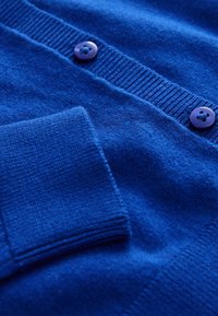 Blauer, gestrickter Cardigan mit gerippten Bündchen und Knopfverschluss, ausgestattet mit violetten Knöpfen und weicher Textur. Nahaufnahme betont die Details des Stoffs.