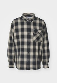 Camisa de franela a cuadros en negro y crema con mangas largas, cierre de botones y un bolsillo en el pecho. Textura suave, diseño con cuello y dobladillo recto.