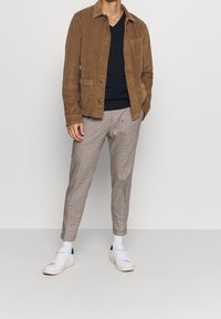 Man som står i en neutral miljö, iförd en brun kordflöjelsjacka, marinblå v-ringad tröja, rutiga byxor och vita sneakers.