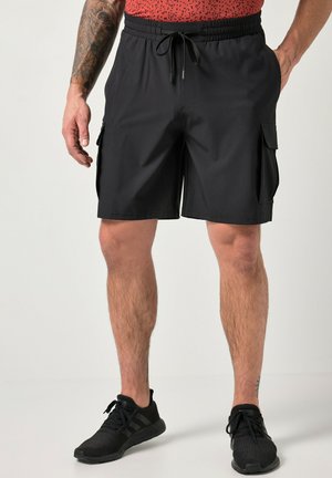 Man draagt zwarte cargoshorts met koord in de taille en zwarte sportschoenen, staat met ontspannen handen langs het lichaam.