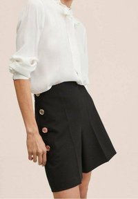 Blusa bianca con colletto arricciato, abbinata a pantaloni corti neri a vita alta con bottoni metallici lucidi sui lati.