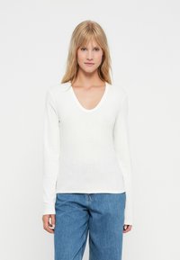 Hvid langærmet top med V-udskæring og ribbet tekstur, parret med blå vide jeans. Har en tæt siddende silhuet.