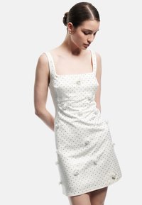 Weißes ärmelloses Kleid mit quadratischem Ausschnitt, verziert mit verstreuten silbernen Nieten und Blumenakzenten, gefertigt aus glattem Stoff.