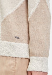 Strickpullover in Beige und Creme, mit einem strukturierten, flauschigen Saum und Bündchen. Silbernes "Bettico"-Etikett auf der linken Seite.