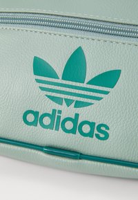 Borsa sintetica verde chiaro con superficie testurizzata, caratterizzata da un logo Adidas color petrolio e un design di foglia a tre strisce sul davanti.