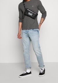 Svart midjeväska med vitt "PUMA"-logotyp, bärs över en grå långärmad tröja, matchad med ljusblå jeans och svarta sneakers med vita detaljer.