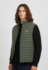 Lightweight green puffer vest with horizontal stitching, high collar, and logo detail. Paired with a black knitted sweater underneath.