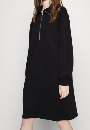 Femme portant une robe noire ample jusqu'aux genoux avec des manches bouffantes longues et un col zippé à l'avant, debout devant un fond uni.