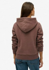 Haut à capuche marron avec une grande capuche, des poignets et un ourlet côtelés. Texture lisse, coupe décontractée. Porté avec un jean en denim bleu, vu de dos.