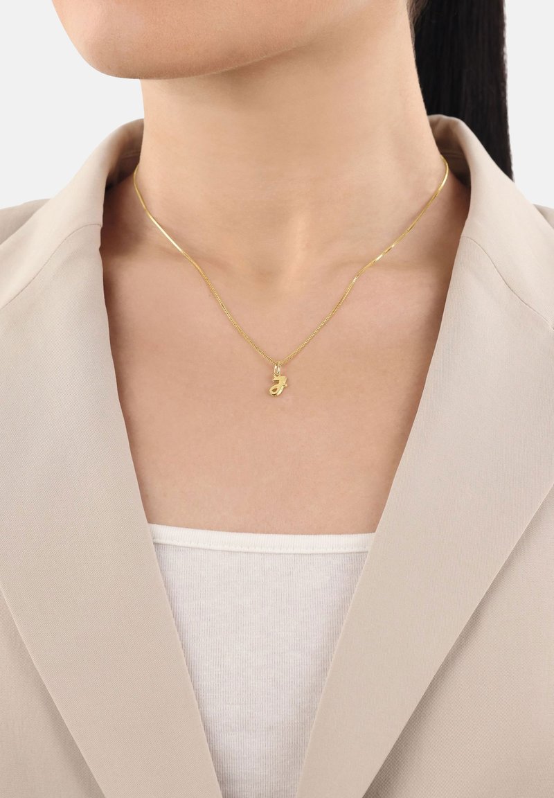 Collar de oro con un pequeño colgante, con una cadena delicada y un acabado brillante, llevado sobre una blusa blanca y debajo de un blazer beige.