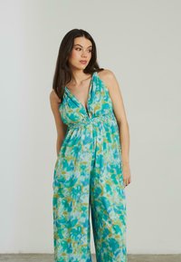 Combinaison fluide en motif tie-dye bleu et vert, avec un décolleté en V profond, un détail de taille tressée et une silhouette à jambes larges. Tissu texturé.