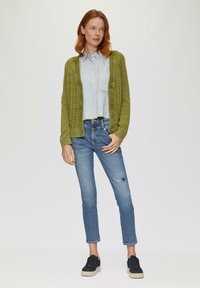 Zielony sweter z dzianiny z wzorem w warkocz, noszony na niebieskiej koszuli w białe paski z kieszonką, w zestawieniu z jasnoniebieskimi dżinsami i czarnymi sneakersami.