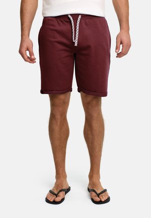 Mann trägt hochgekrempelte dunkelrote Shorts mit weißem Kordelzug, weißes T-Shirt und schwarze Flip-Flops und steht vor einfarbigem Hintergrund.