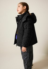 Schwarze Puffjacke mit Kapuze, mit geripptem Stoff und silbernen Druckknöpfen. Getragen über einem blauen Hemd, kombiniert mit dunklen Cargo-Hosen.