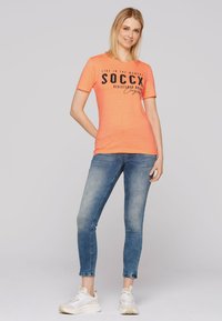 T-shirt a maniche corte di un vivace colore arancione con testo e logo neri. Indossato con jeans blu slim-fit e sneakers bianche. Look semplice e casual.
