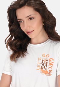 Valkoinen t-paita lyhyillä hihoilla, jossa on graafinen printti ruskeasta ja oranssista cowboy-kengästä sekä teksti "LET'S GO GIRLS" oranssilla.