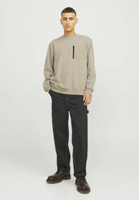 Sweatshirt gris clair avec une poche à rabat olive contrastante, associé à un pantalon cargo noir avec des coutures blanches. Bottes à lacets marron.