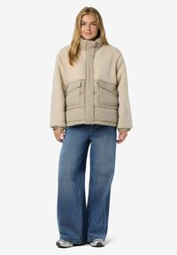 Kobieta ubrana w beżową kurtkę z sherpy i pikowaną, szerokie niebieskie dżinsy oraz srebrne sneakersy, stojąca na tle białym.