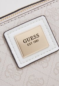 Gros plan sur un tissu beige avec un motif floral subtil, un patch en cuir blanc et une plaque en métal doré gravée avec "GUESS EST 1981" près de la fermeture éclair.