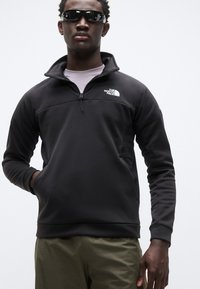 Μαύρο φλις φούτερ με φερμουάρ μέχρι τη μέση, ψηλό γιακά, πλαινά πάνελ από δίχτυ και λογότυπο στο στήθος. Φορεμένο με ελαφριά χακί παντελόνια.