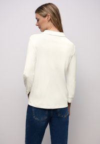 Polo bianca a maniche lunghe con colletto, caratterizzata da una texture liscia e maniche aderenti, abbinata a jeans in denim blu scuro.