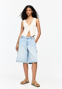 Helles beige geripptes Crop-Top mit V-Ausschnitt und Knopfverschluss, kombiniert mit knielangen hellblauen Jeansshorts und flachen Sandalen.