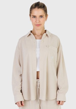 Vrouw met blond haar in een beige oversized blouse over een wit crop top en bijpassende beige broek, staand tegen een witte achtergrond.