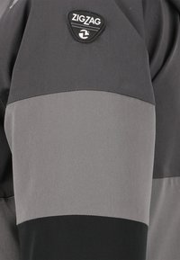 Grå jakkeærme med et zigzag-logo, kontrasterende mørkegrå og sorte sektioner, med glat tekstil og forstærkede syninger.