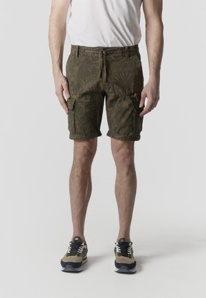 Mężczyzna ubrany w ciemnozielone, wzorzyste szorty bojówki z podwiniętymi nogawkami, białą koszulkę oraz beżowo-czarno-białe sneakersy, stojący na szarym tle.