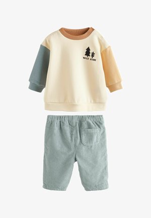 Kermakotona huppari ruskeilla ja vaaleansinisillä hihoilla, jossa on "WILD VIBES" -puugrafiikka; yhdistettynä teal-värisiin corduroy-shortsihousuihin, joissa on tasku.