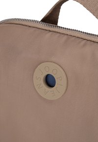 Bolso de tela beige con una superficie texturizada, que presenta una etiqueta redonda beige de la marca y un detalle de botón azul en la parte superior, cerrado con un zip.