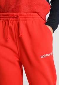 Punaiset verkkarit, valmistettu pehmeästä kankaasta, jossa on joustava vyötärö, kiristettävä nauha ja valkoinen Adidas-logo vasemmassa reidessä.