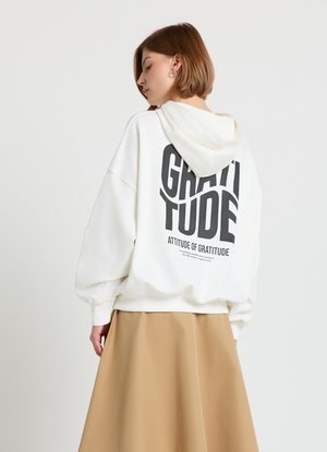 Weißes Kapuzen-Sweatshirt mit großem schwarzem Schriftzug "GRATITUDE" auf dem Rücken. Dazu ein beiger Rock mit entspanntem Schnitt und weicher Textur.