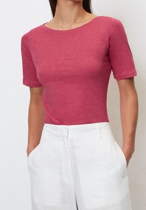 Frau trägt ein kurzärmeliges, figurbetontes, dunkelrosa Oberteil, das in eine hoch taillierte weiße Hose gesteckt ist, mit Händen in den Taschen.