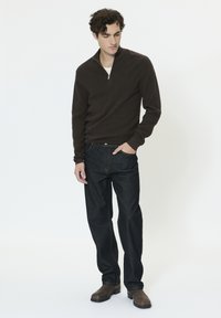 Jeune homme portant un pull à col zippé marron foncé, un jean en denim foncé et des bottes en cuir marron, debout avec une main dans la poche.