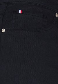 Tissu en denim noir mettant en valeur une poche avant avec un petit emblème tricolore et un bouton en argent. La texture est lisse avec un léger éclat.