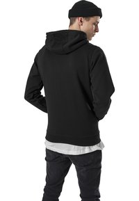 Sudadera negra con capucha forrada, dobladillo y puños acanalados, llevada sobre una camiseta blanca, combinada con unos vaqueros oscuros, vista desde la parte posterior.