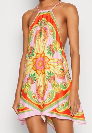 Kobieta ubrana w kolorową sukienkę na szyję z kwiatowymi i paisley wzorami w kolorach różu, pomarańczu, żółci i zieleni oraz asymetrycznym dołem.