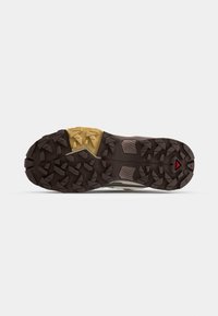 Semelle extérieure robuste en caoutchouc marron foncé et beige d'une chaussure de trail, avec des crampons profonds multidirectionnels pour la traction.