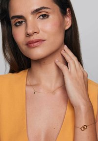 Złoty łańcuszek z zawieszką w kształcie kwiatu i zielonym akcentem, w zestawie ze złotą bransoletką z wzorem splecionych pierścieni. Widoczne odcienie i tekstury skóry.