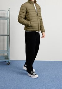 Olivgrön dunjacka med horisontella sömndetaljer, kombinerad med svarta manchesterbyxor och svartvita sneakers. Blå texturerad golv.