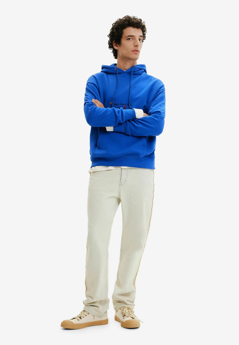 Niebieska bluza z kapturem z przednią kieszenią, białymi mankietami i dekoracyjnym haftem, w połączeniu z jasnobeżowymi dżinsami i beżowymi sneakersami.