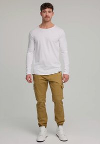 Jeune homme debout, portant une chemise blanche à manches longues, un pantalon cargo beige et des baskets blanches, sur un fond neutre.