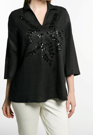 Blouse noire à col en V et manches trois-quarts avec des motifs de feuilles en sequins noirs brillants, associée à un pantalon blanc.
