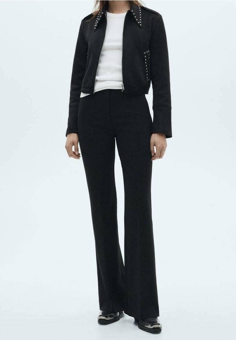 Femme portant une veste noire cloutée, une chemise blanche, un pantalon noir taille haute évasé et des chaussures noires sur un fond uni.