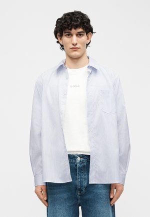 Jeune homme aux cheveux bouclés portant un T-shirt blanc à motif sous une chemise boutonnée bleu clair à rayures ouverte et un jean bleu.