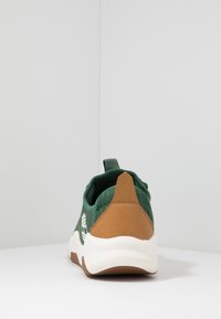 Chaussure de sport en tissu vert avec un capuchon de talon en cuir brun, semelle blanche et un motif de logo décoratif sur le côté.