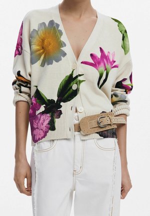 Kvinde iført beige cardigan med store farverige blomstertryk, et beige bælte med nitter og højtaljede hvide bukser med synlige sømme.