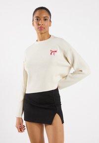 pulover crem cu model striat, având un detaliu brodat sub formă de fundiță roz pe partea stângă a pieptului, asociat cu o fustă mini neagră cu o tăietură laterală.